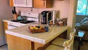 A kitchen with wooden cabinets, a gas stove, a coffee maker, and a small dining area with a white chair. - Auburn Party of 8 (Auburn)