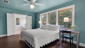 A bedroom with a bed, a desk, a chair, a window with a view, and a ceiling fan.