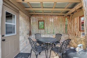 A covered patio area with stone walls, a wooden ceiling, a window, and a round table with chairs. - Milk Bath Springs | Pet-Friendly Historic Downtown Cottage (Dripping Springs)