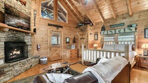 Un interior de cabaña acogedor con chimenea de piedra, paredes de madera, un sofá de cuero cómodo y una cama con manta blanca.