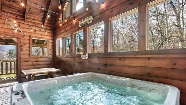 Una cabaña de madera con jacuzzi, paredes de madera, ventanas grandes y luces colgantes.