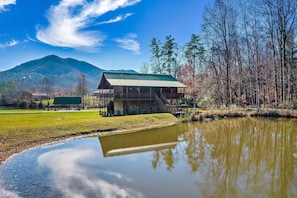 A cabin with a green roof and a wooden deck, surrounded by a pond and trees, with a mountain in the background.