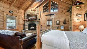 Interior de una cabaña de troncos con chimenea de piedra, una cama, un sofá de cuero y techo de madera.