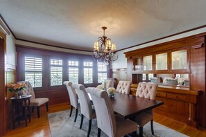 A dining room with a large wooden table, six upholstered chairs, a chandelier, and a kitchen area with wooden cabinets and a window. - Cozy 1920's Craftsman bungalow, Hillcrest/Mission Hills (San Diego)
