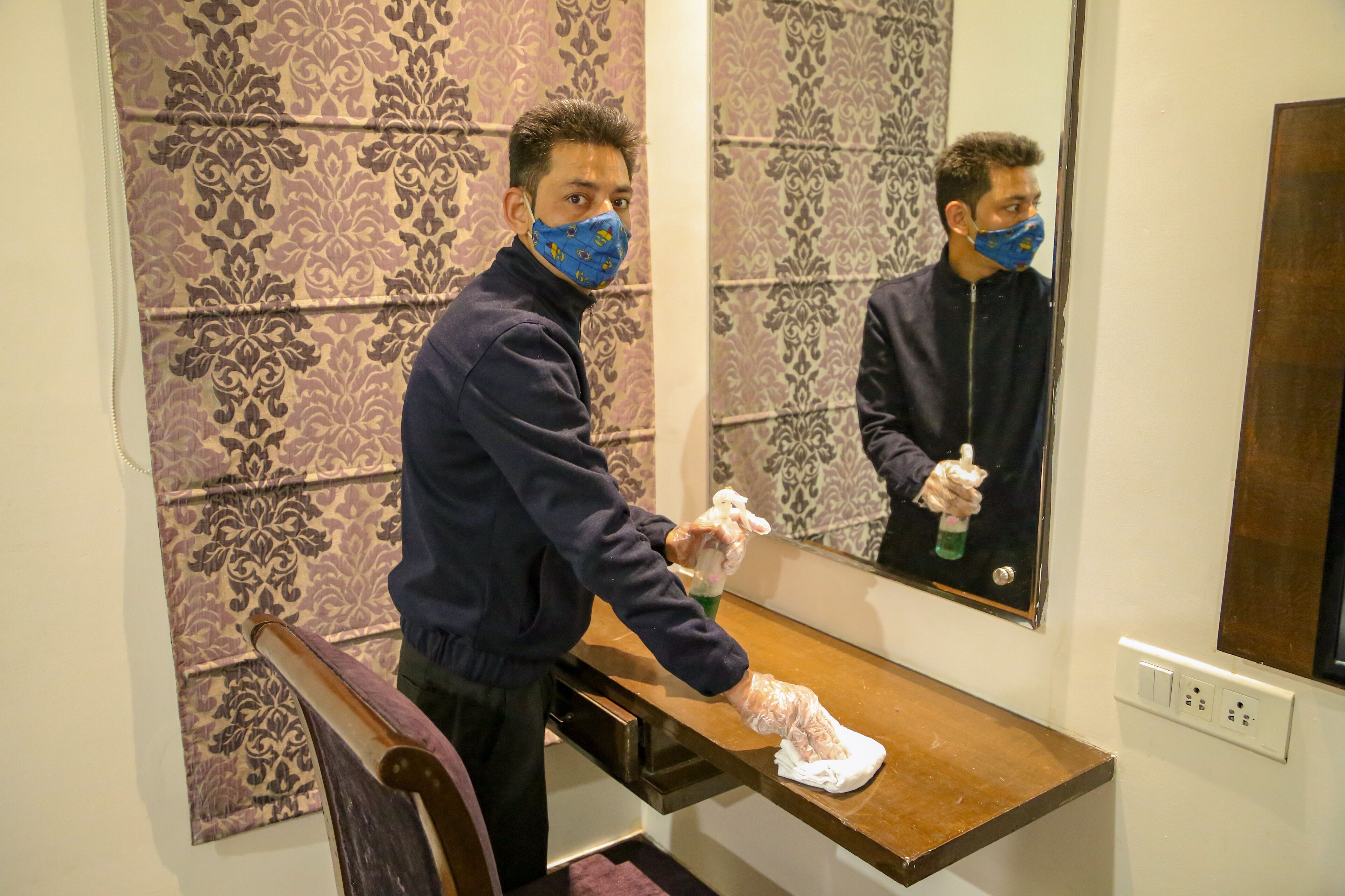 A person cleaning a wooden counter in a room with patterned wallpaper and a mirror reflecting the scene.