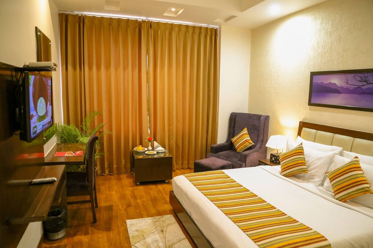 A hotel room with a bed, a desk with a television, a chair, a nightstand with a lamp, and a window with curtains.