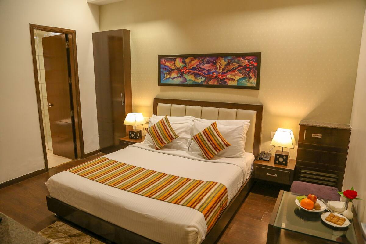 A hotel room with a bed, bedside tables, a nightstand, a dresser, a painting, and a glass table with fruit and a vase.