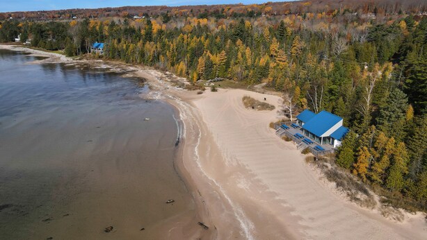 Una casa de playa con techo azul, rodeada de árboles de colores otoñales y una playa arenosa.