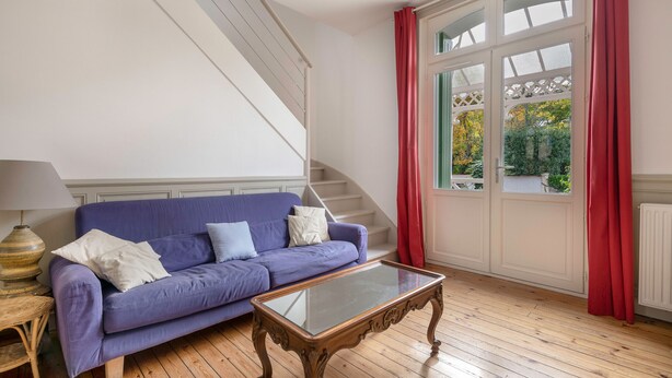 Una habitación con un sofá púrpura, una mesa de centro de cristal, una escalera de madera y un ventanal con cortinas rojas.