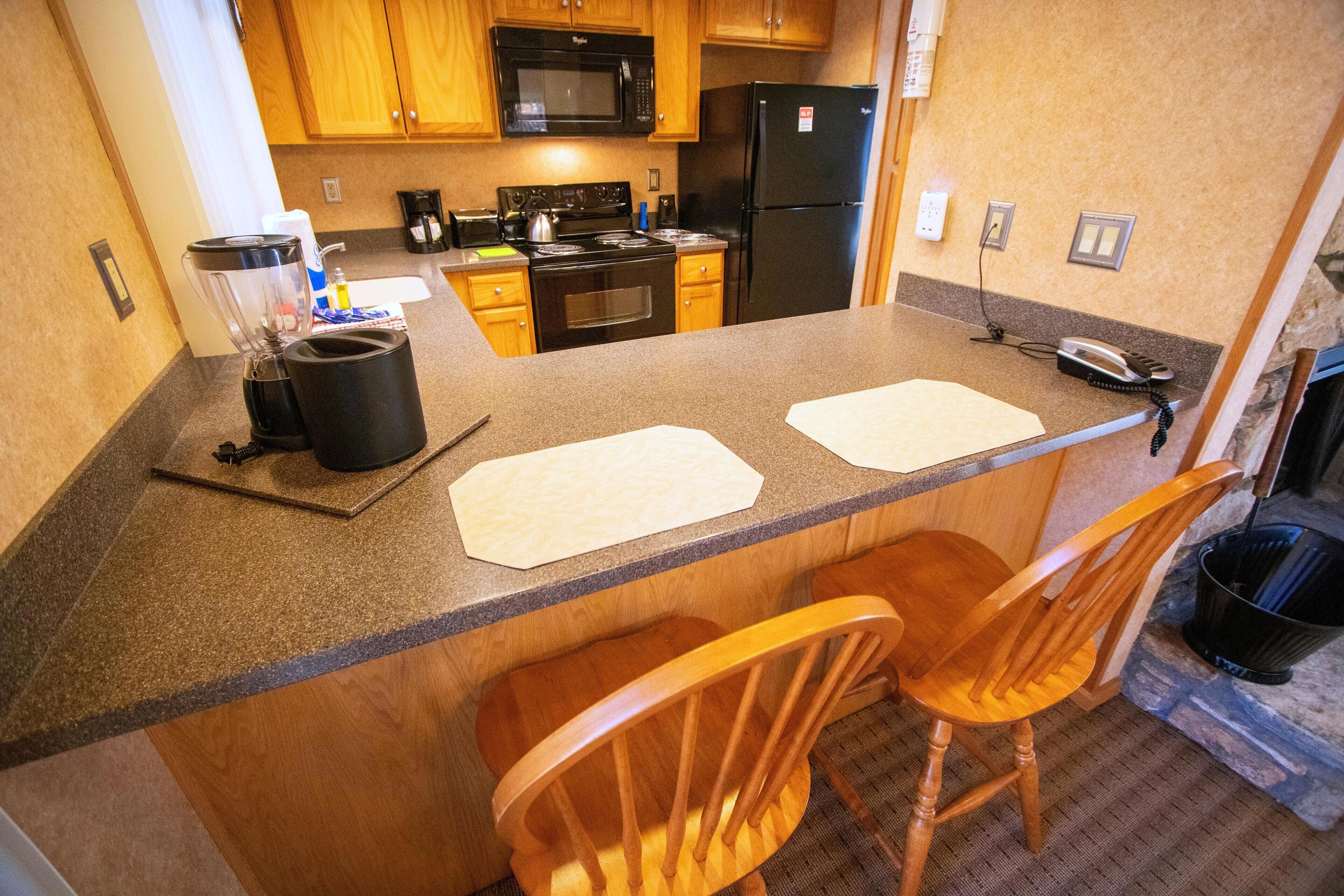 A compact kitchen with wooden cabinets, a black refrigerator, a microwave, and a stove. There are two wooden chairs and a bar stool.