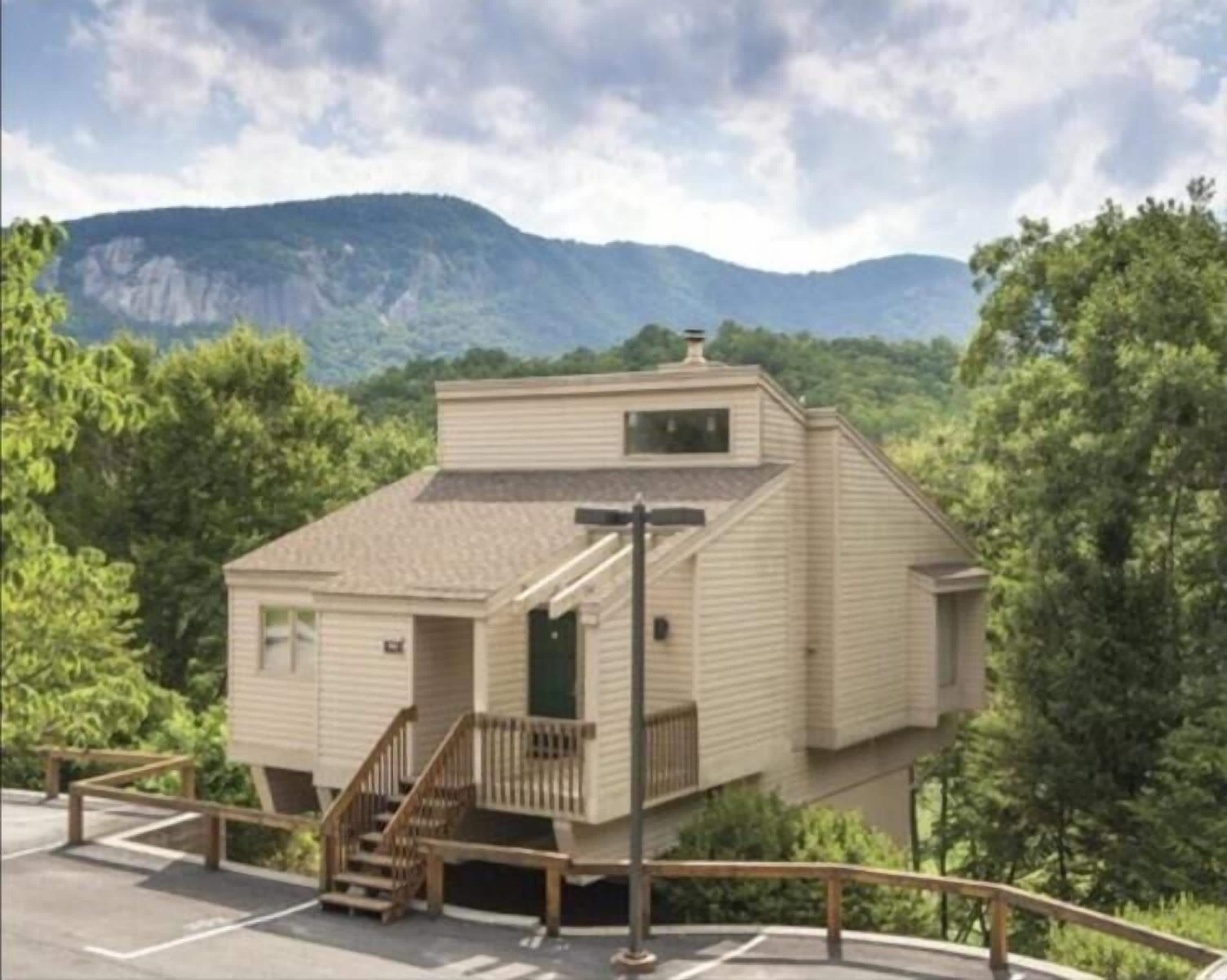 A two-story cabin with a small porch and a staircase, surrounded by trees and mountains.