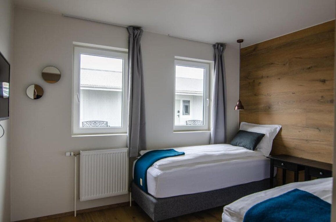 A bedroom with two beds, a wooden headboard, a radiator, and two windows with curtains.
