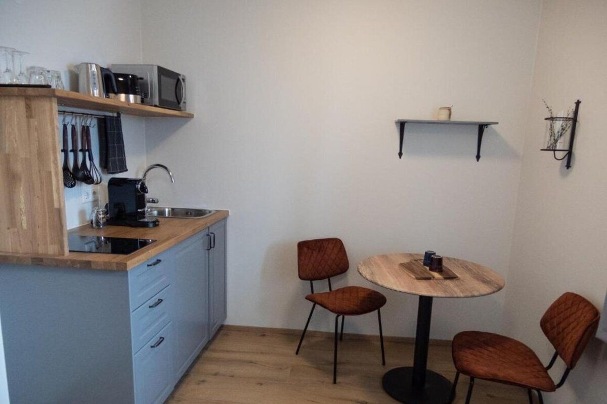 Una cocina compacta con microondas, tostadora y lavabo, una pequeña mesa de comedor con cuatro sillas, y un estante con una taza y un objeto decorativo.