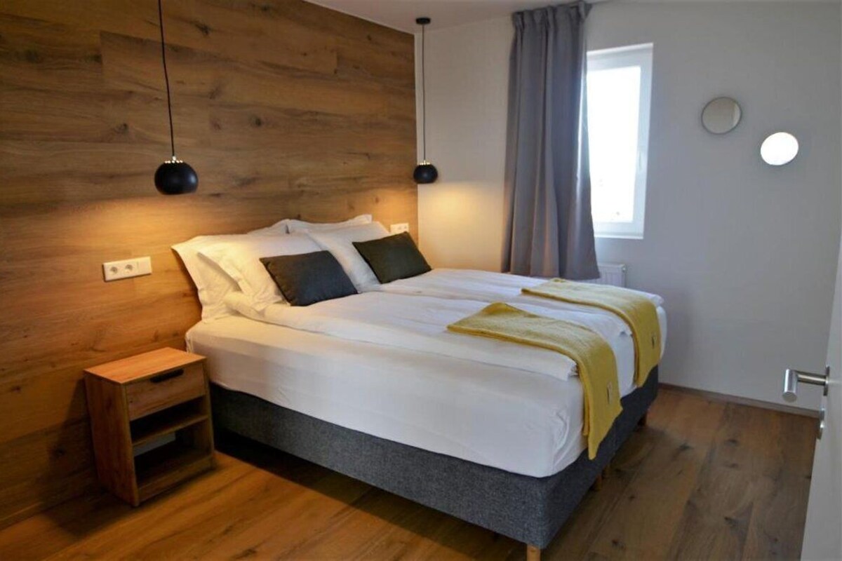 Un dormitorio con cabecera de madera, cama con ropa de cama blanca y una manta amarilla, mesita de noche y ventana con cortinas.