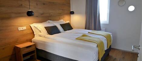 A bedroom with a wooden headboard, a bed with white bedding, two bedside lamps, a bedside table, and a window with curtains.
