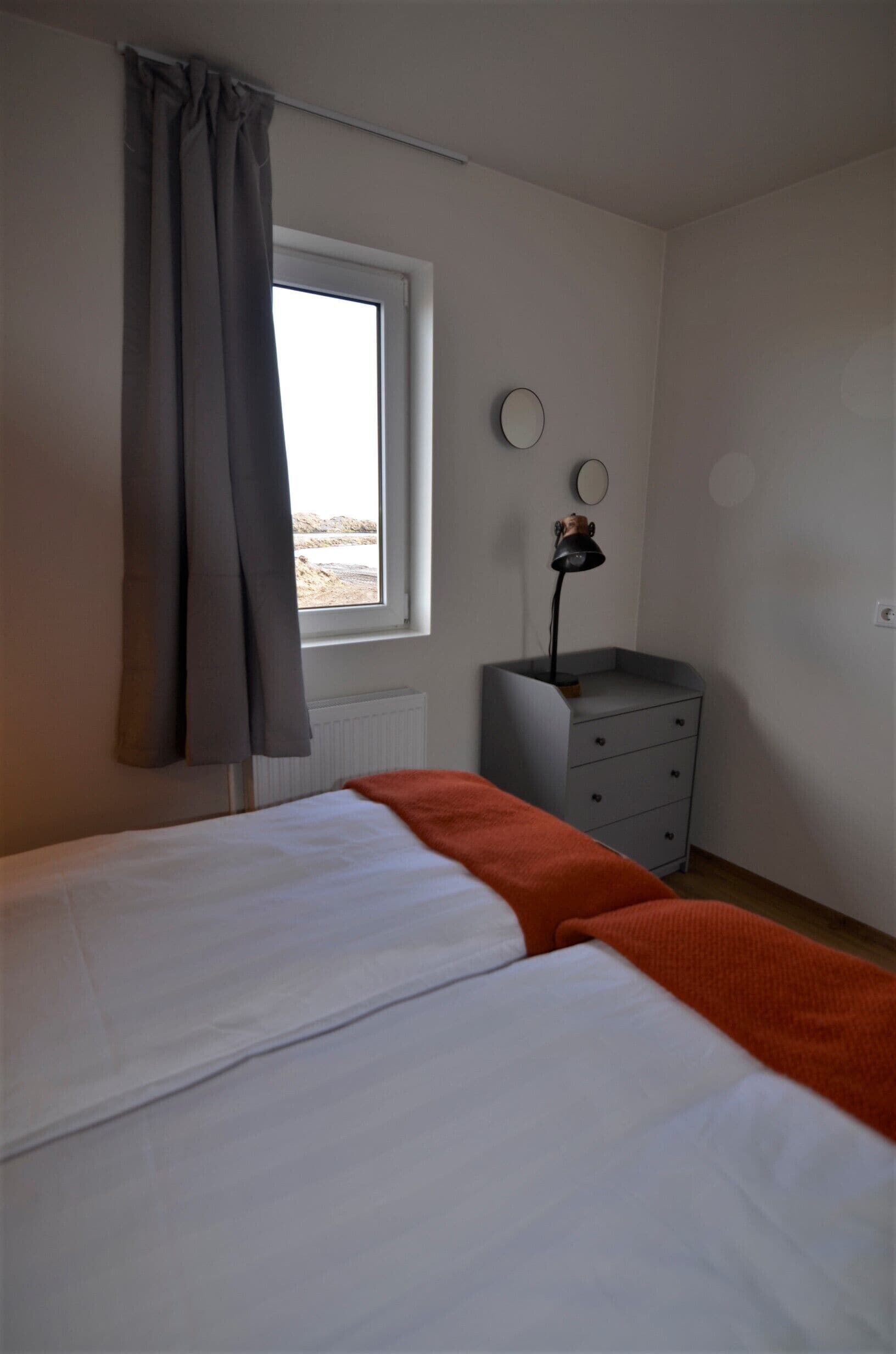 A bedroom with a bed, a window with curtains, a bedside table with a lamp, and a dresser.