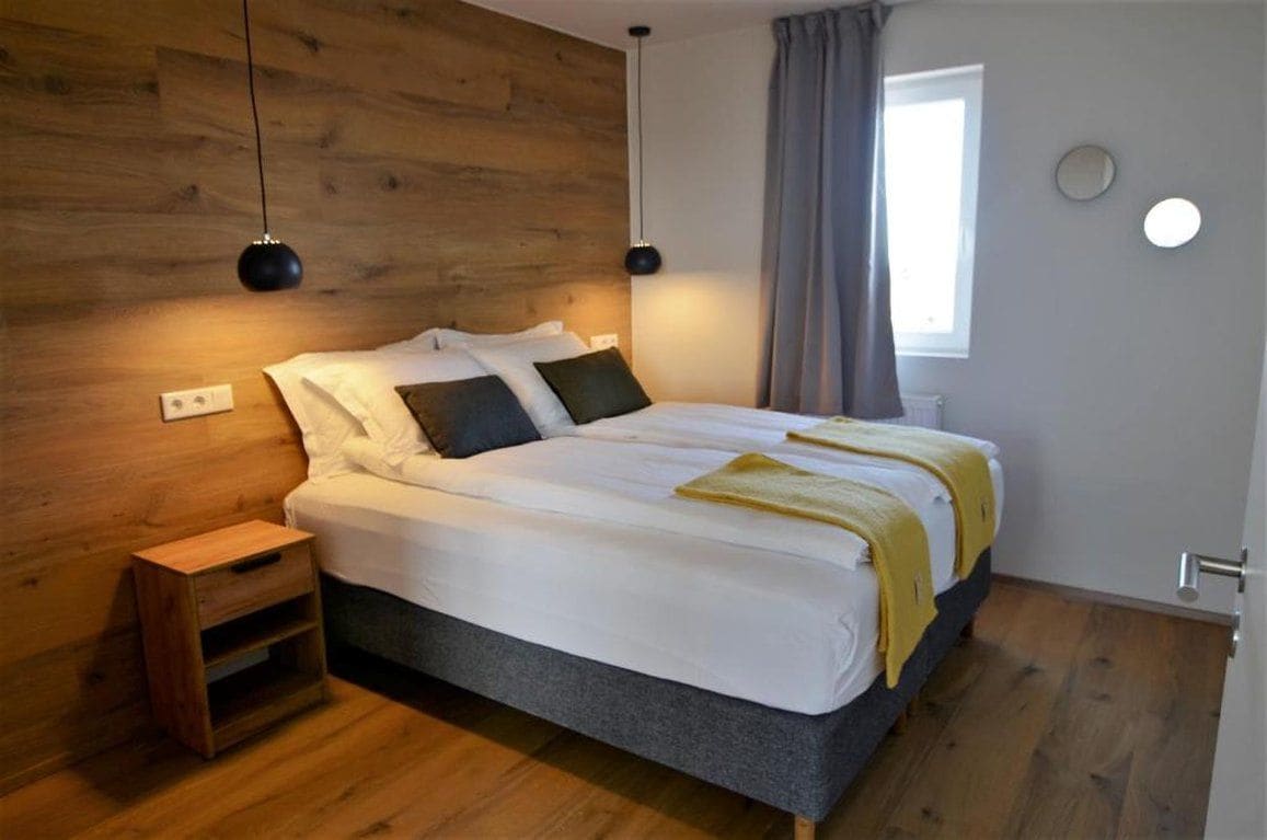 A bedroom with a wooden headboard, a bed with white bedding, two bedside lamps, a bedside table, and a window with curtains.