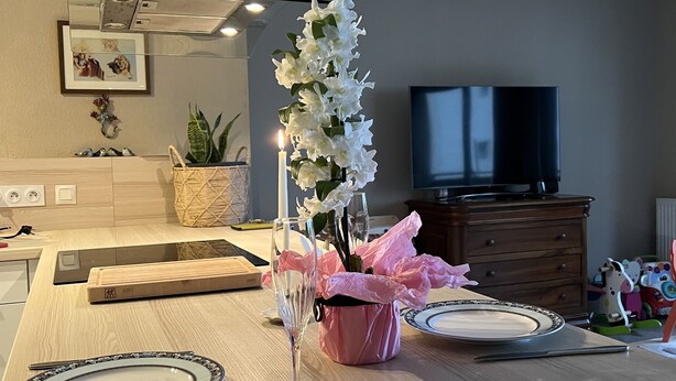 Une table à manger est dressée avec des assiettes, des couverts et un verre d’eau. Un vase rempli de fleurs blanches et une boîte de mouchoirs rose sont posés sur la table.