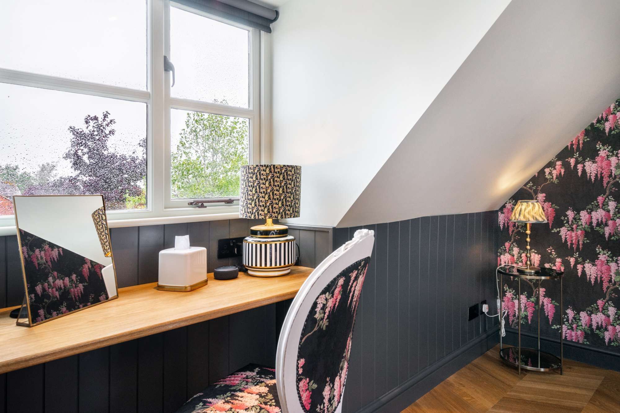 A modern loft bedroom with a sloped ceiling, a wooden desk, a chair, a lamp, and a wall with floral wallpaper.