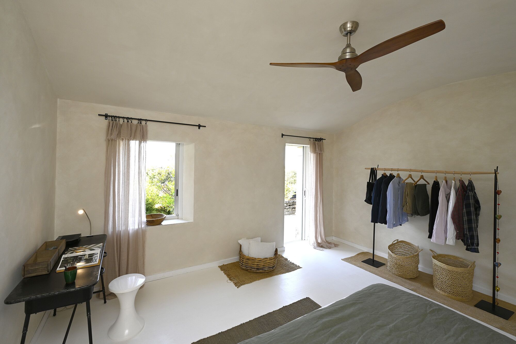 A bedroom with a ceiling fan, a bed, a desk with a chair, a window with curtains, a clothes rack, and a small table with a bowl and a cup.