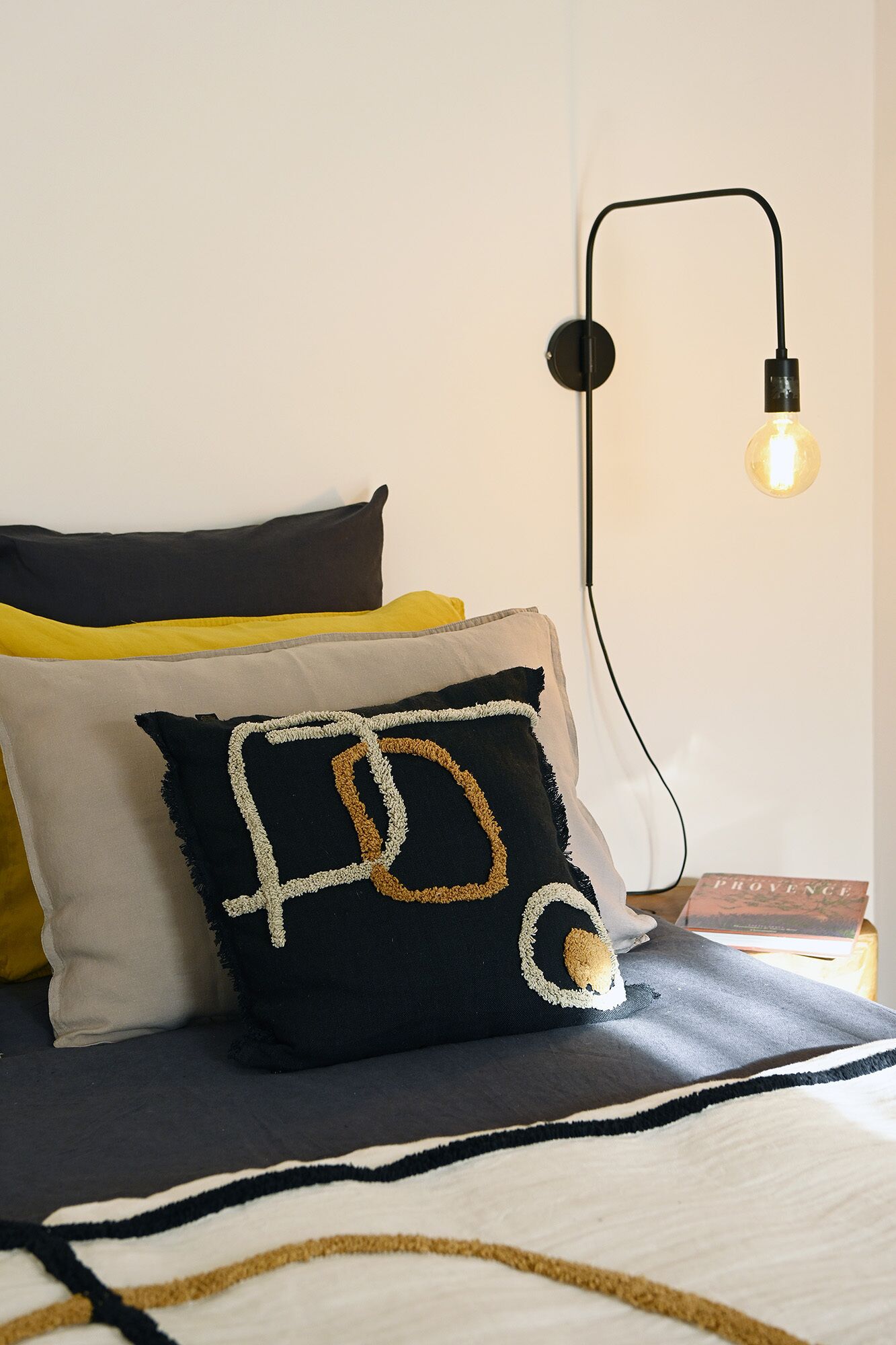 Ein ordentlich gemachtes Bett mit einer schwarz-weiß gemusterten Tagesdecke, einem schwarzen Kissen mit Golddekor und einem gelben Kissen. Eine schwarze Wandlampe mit einer Glühbirne ist sichtbar.