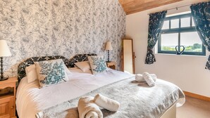 Ein Schlafzimmer mit einem großen Bett, blumigem Wandpapier, einer Holzbalkendecke und einem Fenster mit Vorhängen.