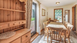 Une salle à manger en bois avec une table et des chaises, un buffet en bois avec des tiroirs, et une vue sur l’extérieur à travers une porte vitrée.