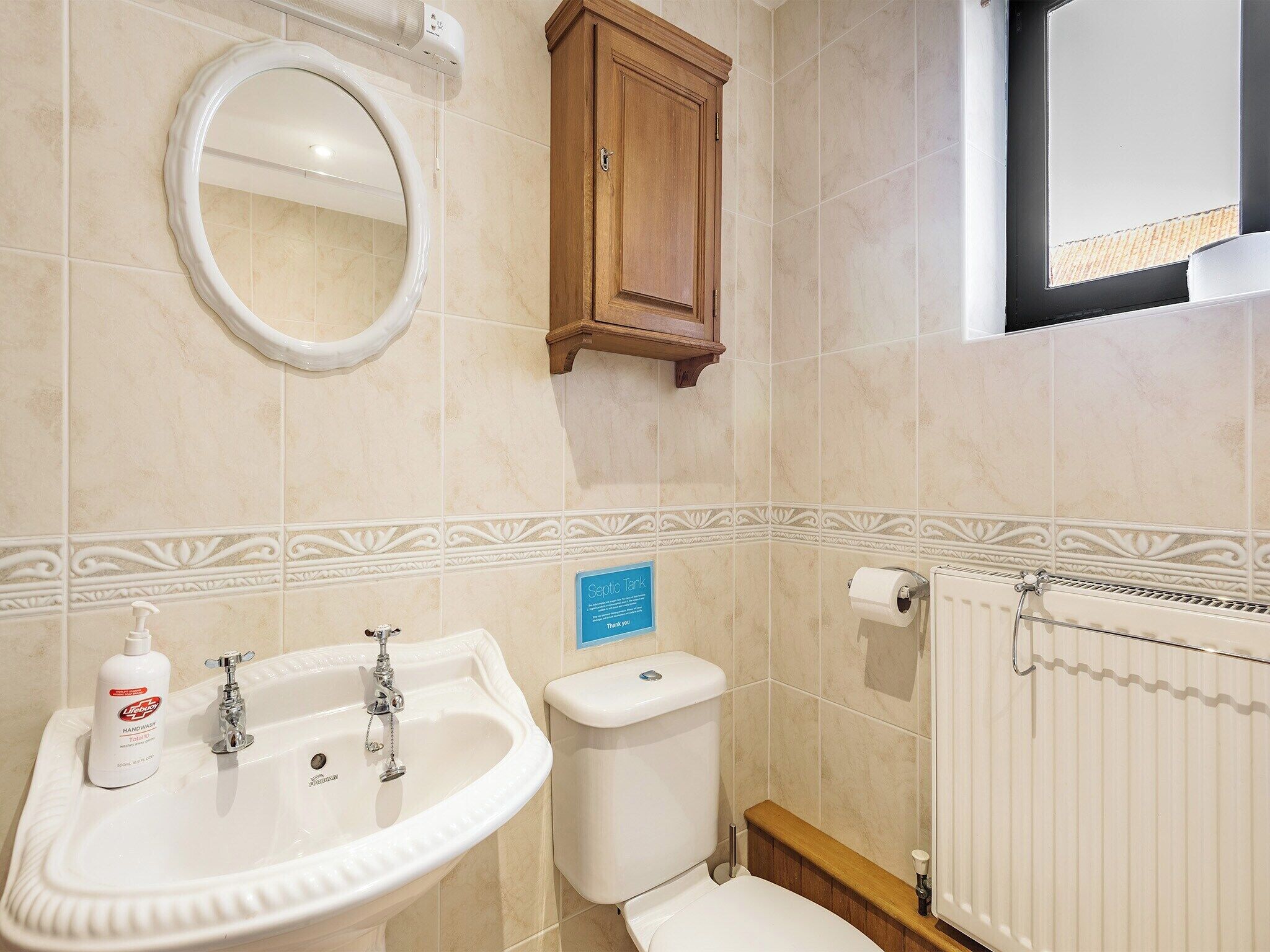 Un baño con lavabo, inodoro y espejo. Hay un armario de madera sobre el inodoro y un cartel en la pared.