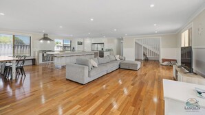 Amplia sala de estar con sofá seccional gris, piso de madera, cocina con armarios blancos y una escalera que sube a un altillo.