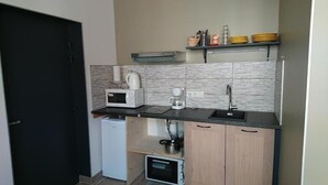 Eine kompakte Küche mit Mikrowelle, Kaffeemaschine und kleinem Backofen. Ein schwarzes Spülbecken und ein Regal mit Geschirr und Tassen.