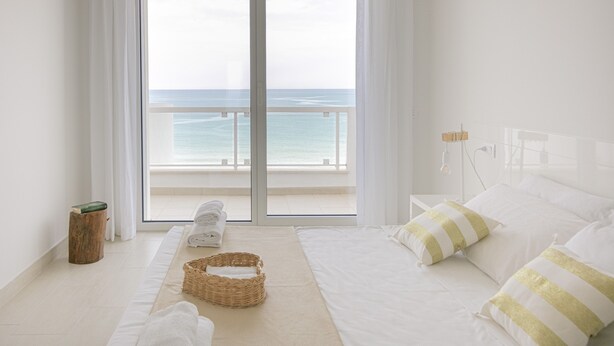 A hotel room with a large window overlooking the sea, a bed with white bedding and striped pillows, a wicker basket, and a small wooden stool with a book on it. - Rivazzurra Homes - 28 (Termoli)