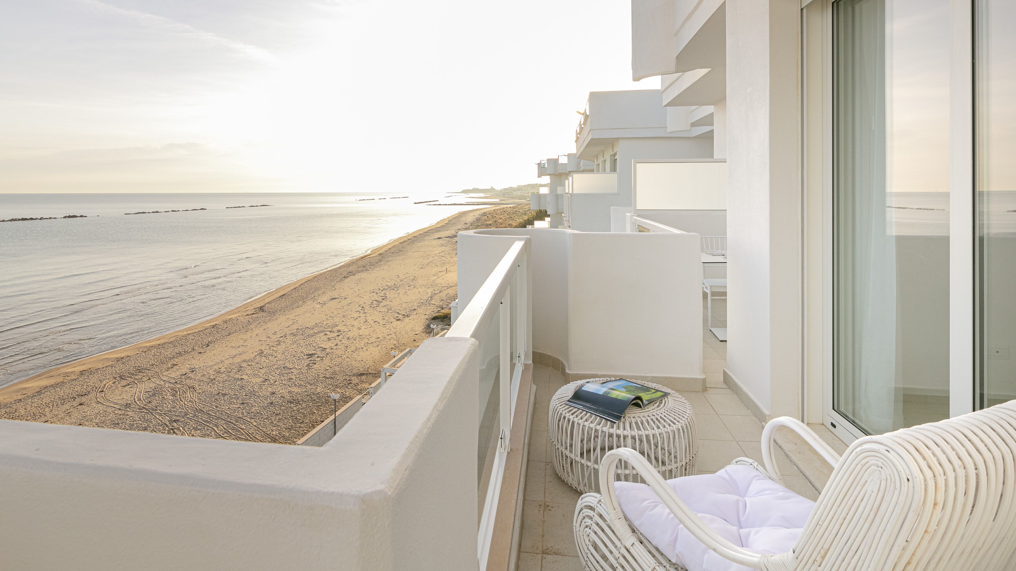 A balcony with a view of the beach, white wicker furniture, and a small table with a book.