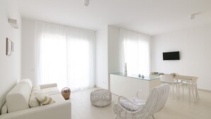 A modern, minimalist living room with a sofa, a wicker chair, a glass-top dining table, and a TV mounted on the wall. - Rivazzurra Homes - 28 (Termoli)