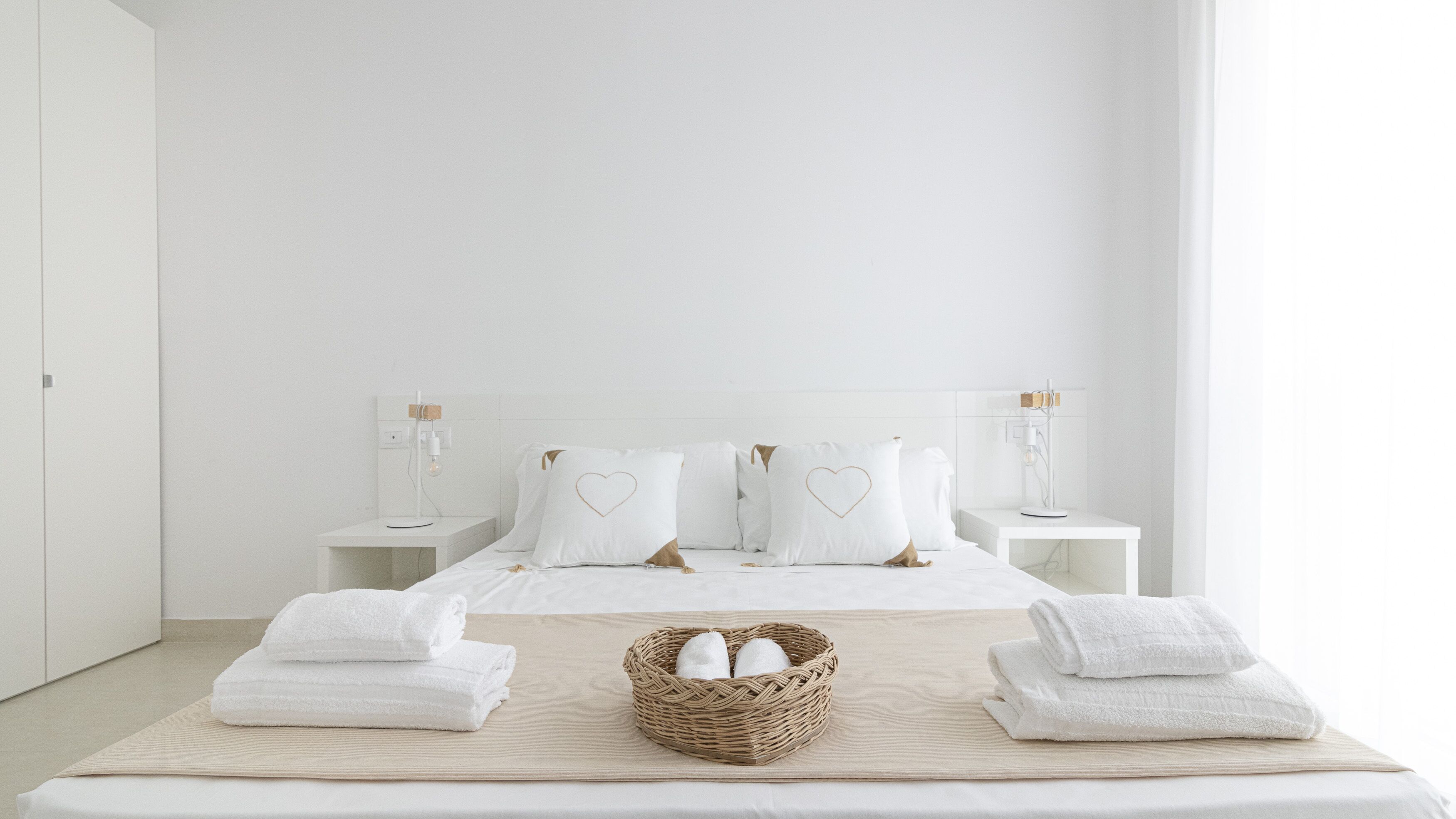 Een minimalistisch hotelkamer met een groot bed, twee nachtkastjes, een bedlamp en een geweven mand met handdoeken.