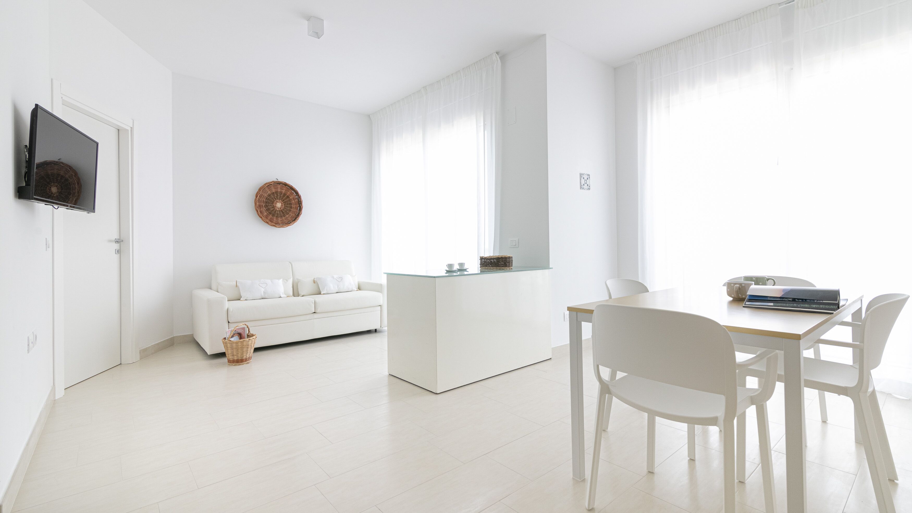Een moderne, minimalistische woonkamer met een witte bank, een salontafel met glazen blad, en een eethoek met witte stoelen.