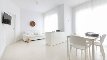 Een moderne, minimalistische woonkamer met een witte bank, een salontafel met glazen blad, en een eethoek met witte stoelen.