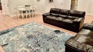 Uma sala de estar com um sofá de couro marrom escuro, um tapete estampado, uma área de jantar com cadeiras brancas e uma mesa, e uma porta com uma guirlanda decorativa.
