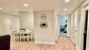 Um corredor amplo com piso de madeira, uma área de jantar com cadeiras brancas e uma mesa, e um luminário fixado na parede.