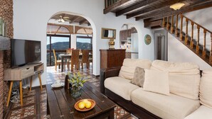 Amplia sala de estar con sofá, TV y mesa de comedor con sillas. Se observa una escalera con barandilla de madera y vista al exterior.