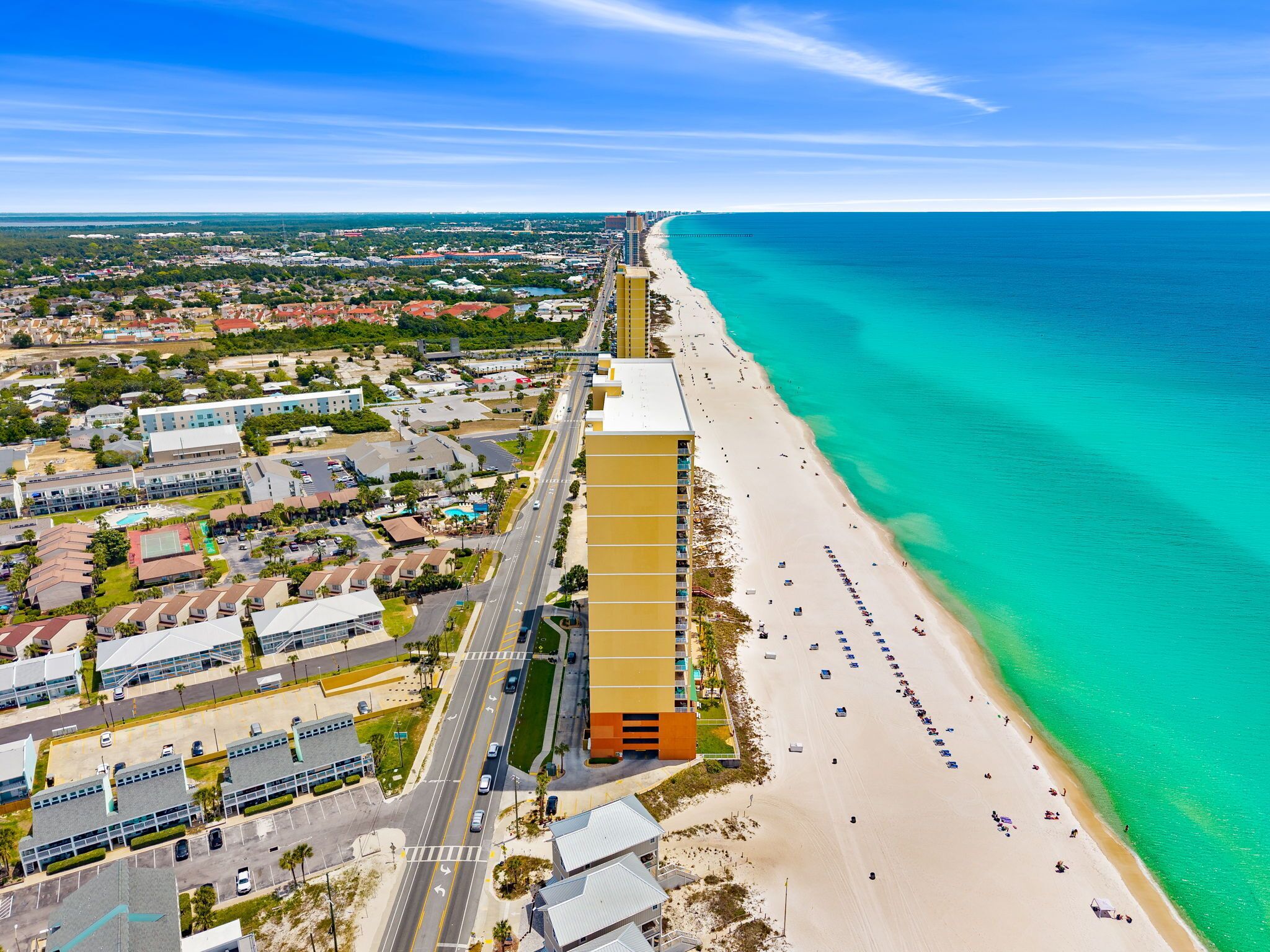 An aerial view of a coastal city with a prominent beach, a long road parallel to the shore, and a large building complex with multiple floors.