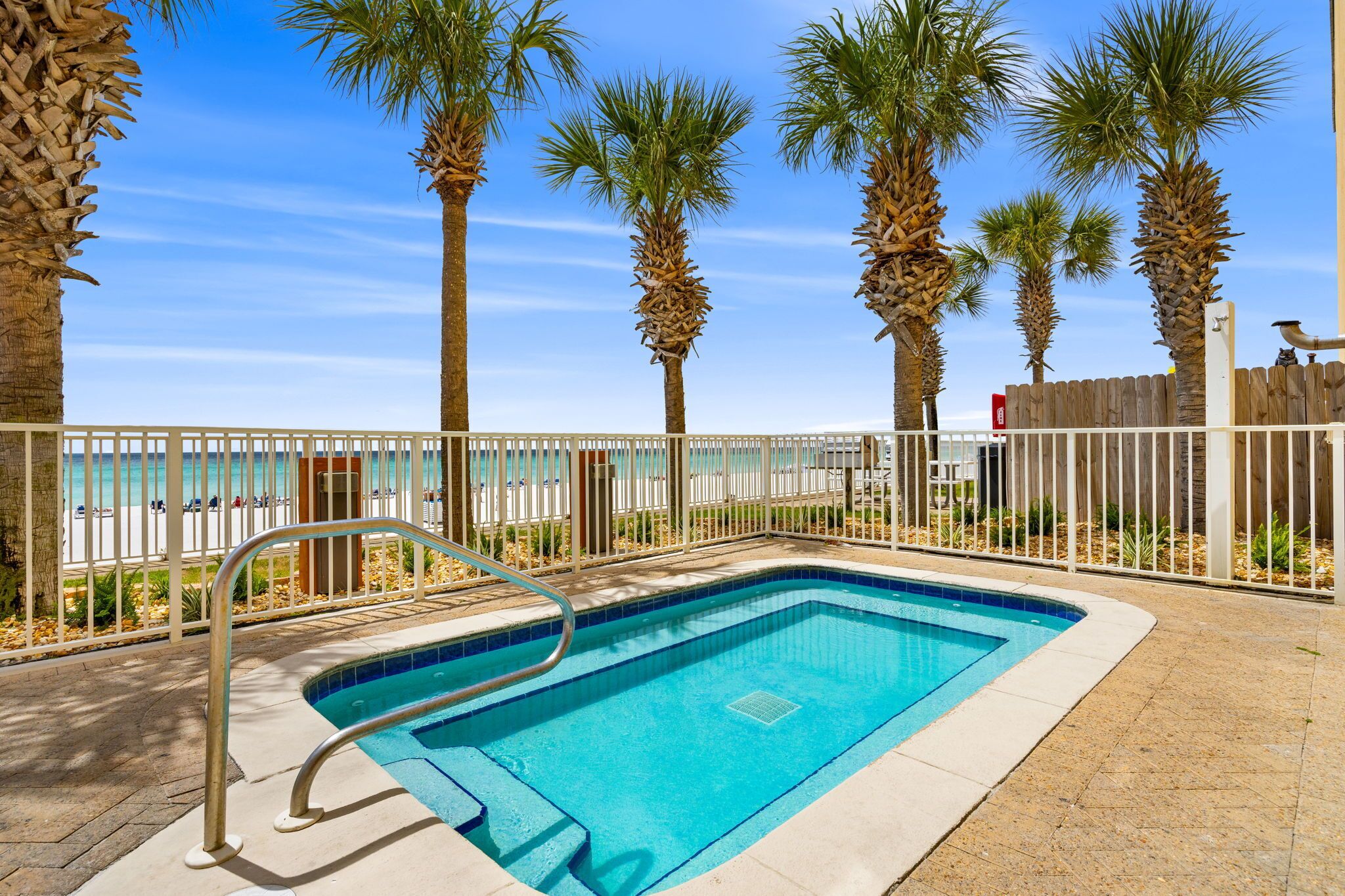 A rectangular pool with a metal ladder, surrounded by a white fence, palm trees, and a view of a beach.