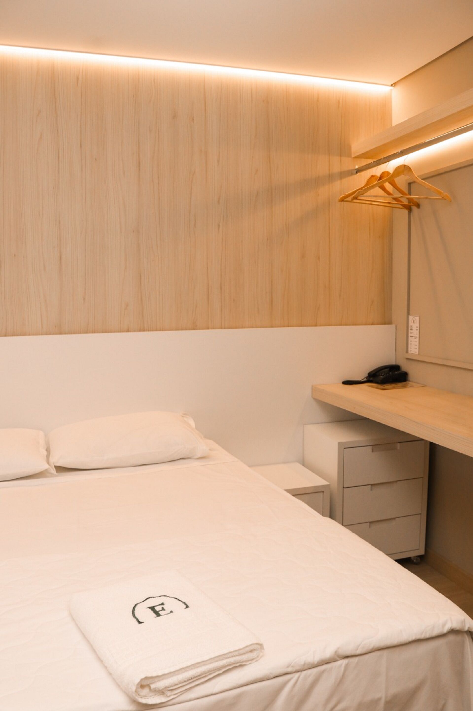 A hotel room with a neatly made bed, a bedside table with a phone, a hanging clothes rack, and a small desk with drawers.