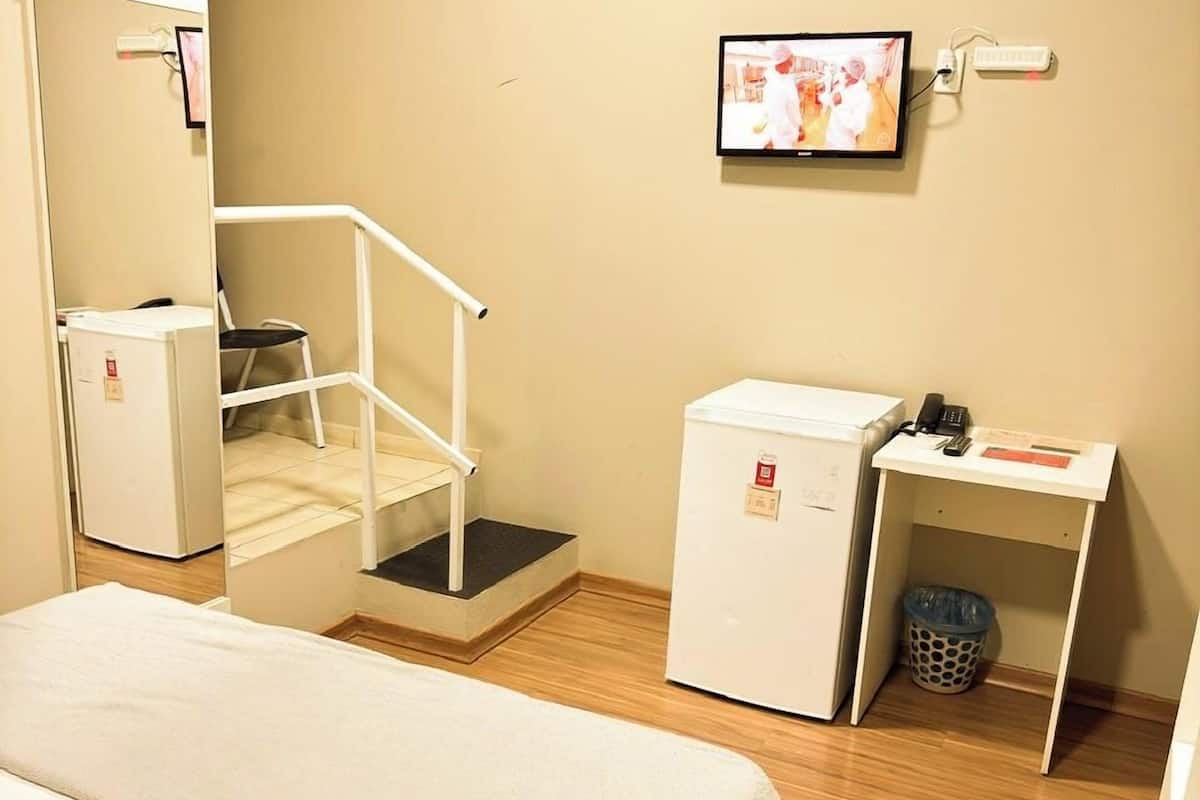 A hotel room with a bed, a desk, a chair, a trash bin, and an air conditioning unit.
