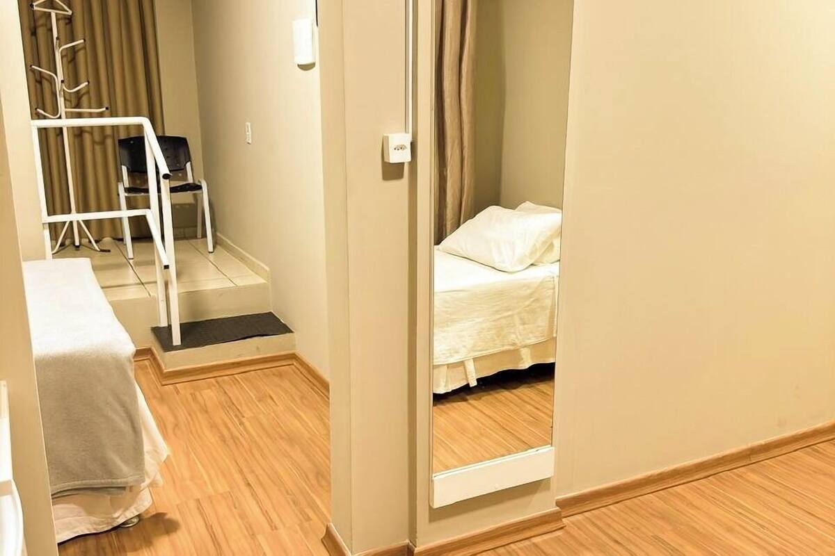 A hotel room with a wooden floor, a bed with white bedding, a chair, a desk, and an air conditioning unit.