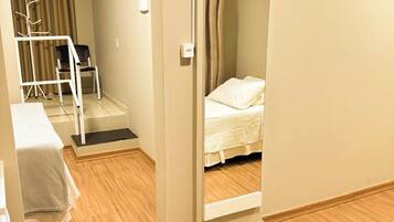 Una habitación de hotel con piso de madera, una cama con ropa de cama blanca, una silla, un escritorio y un equipo de aire acondicionado.