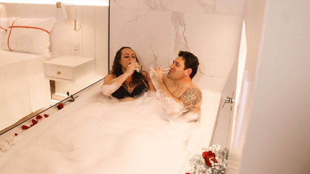 A couple in a bathtub with a white marble wall and a small white cabinet. - Estação Termas Hotel (Piratuba)