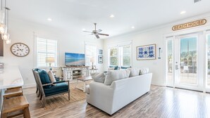 A spacious living room with a white sofa, a blue armchair, and a wooden coffee table. There is a ceiling fan, a clock, and a sign that reads "BEACH HOUSE". - Walk to the Beach ~ Golf Cart ~ Communal Pool (Santa Rosa Beach)