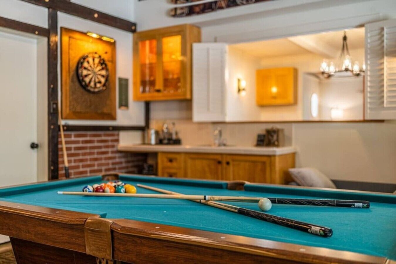 A pool table with balls and cues, a dartboard, wooden cabinets, a brick wall, and a chandelier.