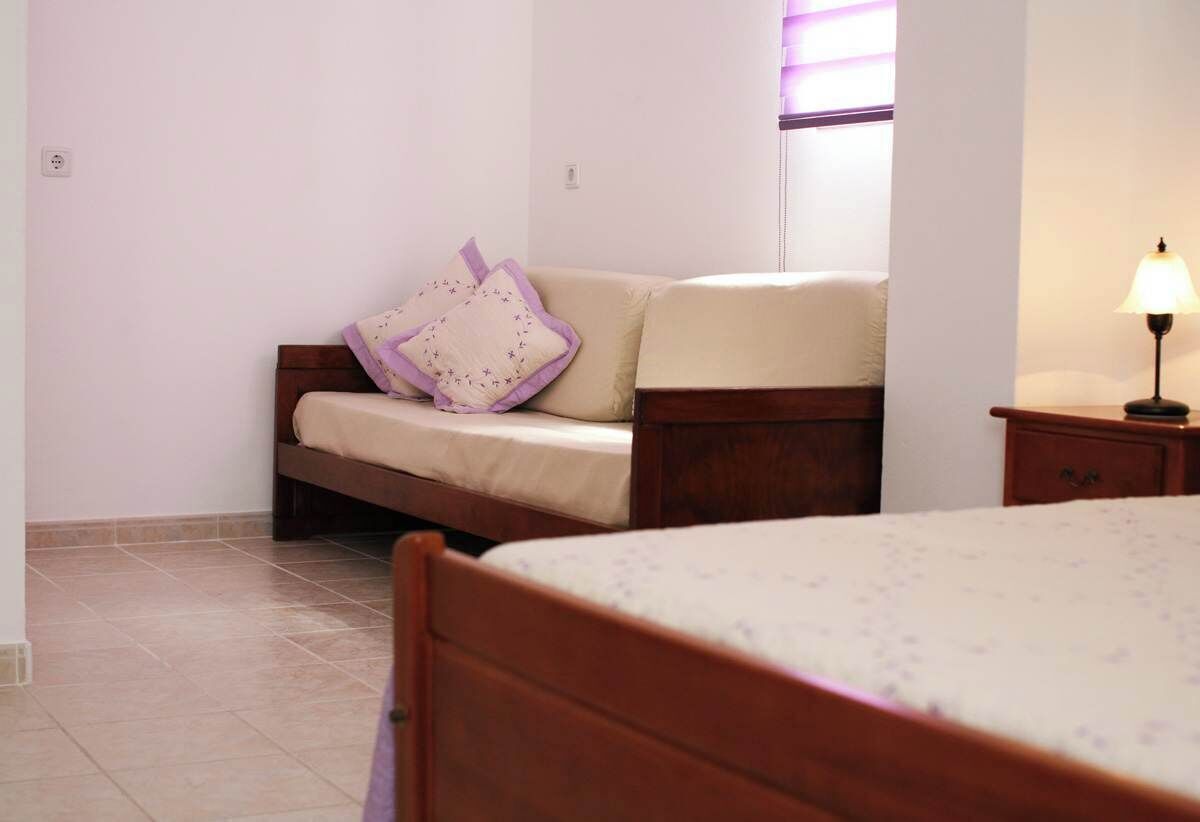 A bedroom with a bed, a wooden nightstand with a lamp, a sofa with cushions, and a window with blinds.