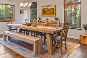 A dining area with a wooden table, benches, and chairs, large windows with a view of trees, and a chandelier.